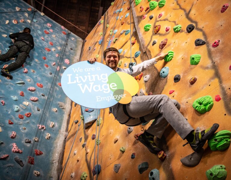 Steven Rome on a climbing wall with a living wage logoboard