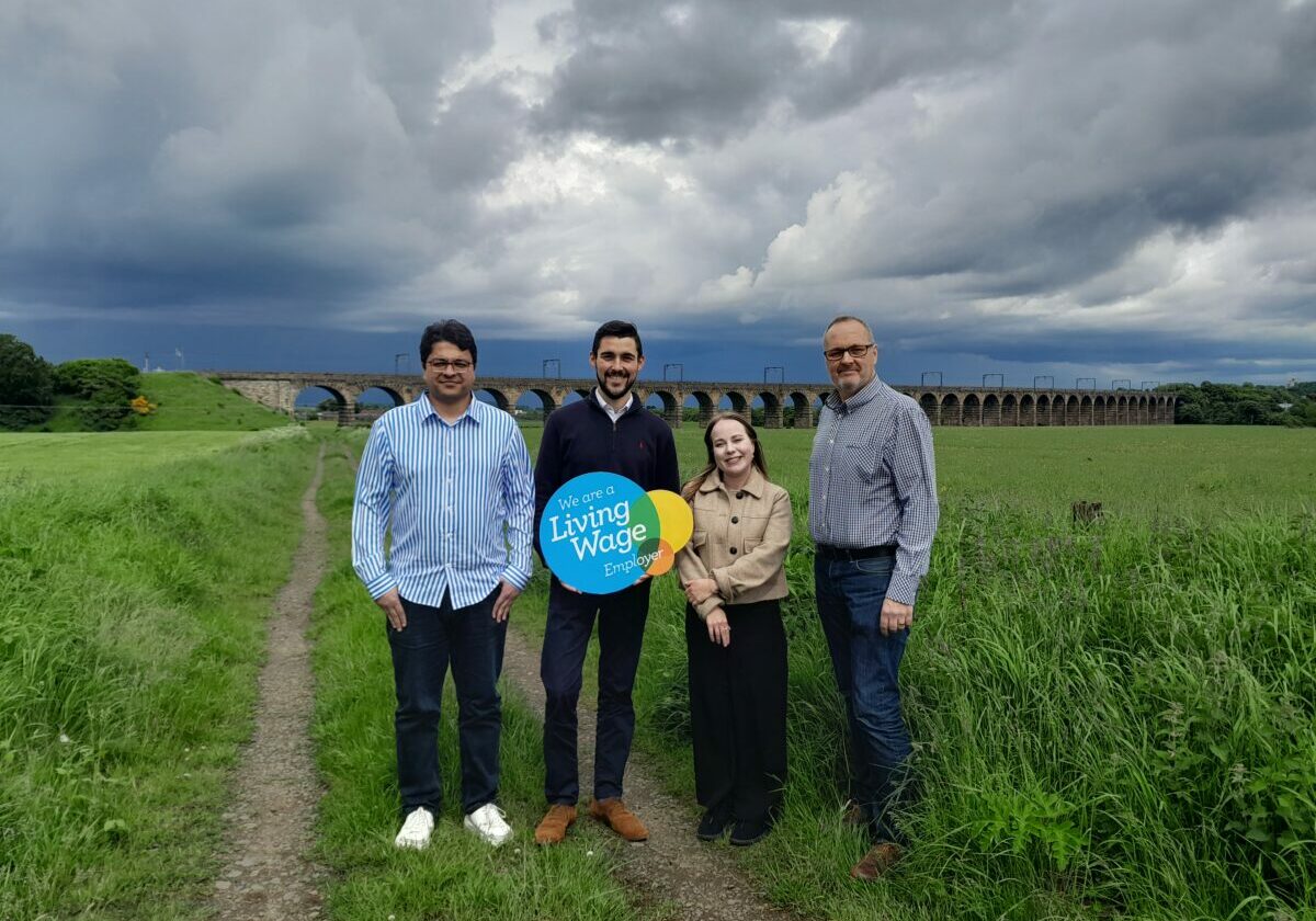 Four people stand in front of farm land, holding a Living Wage Scotland Logoboard