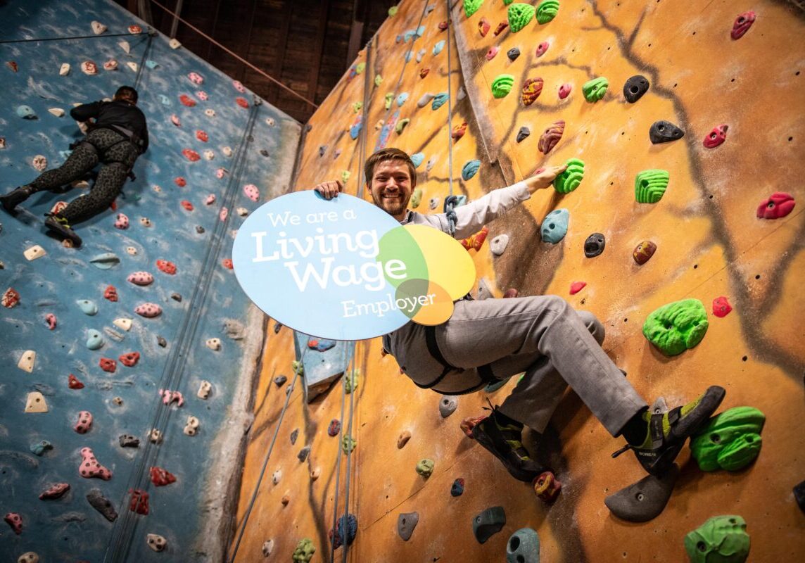 Steven Rome on a climbing wall with a living wage logoboard