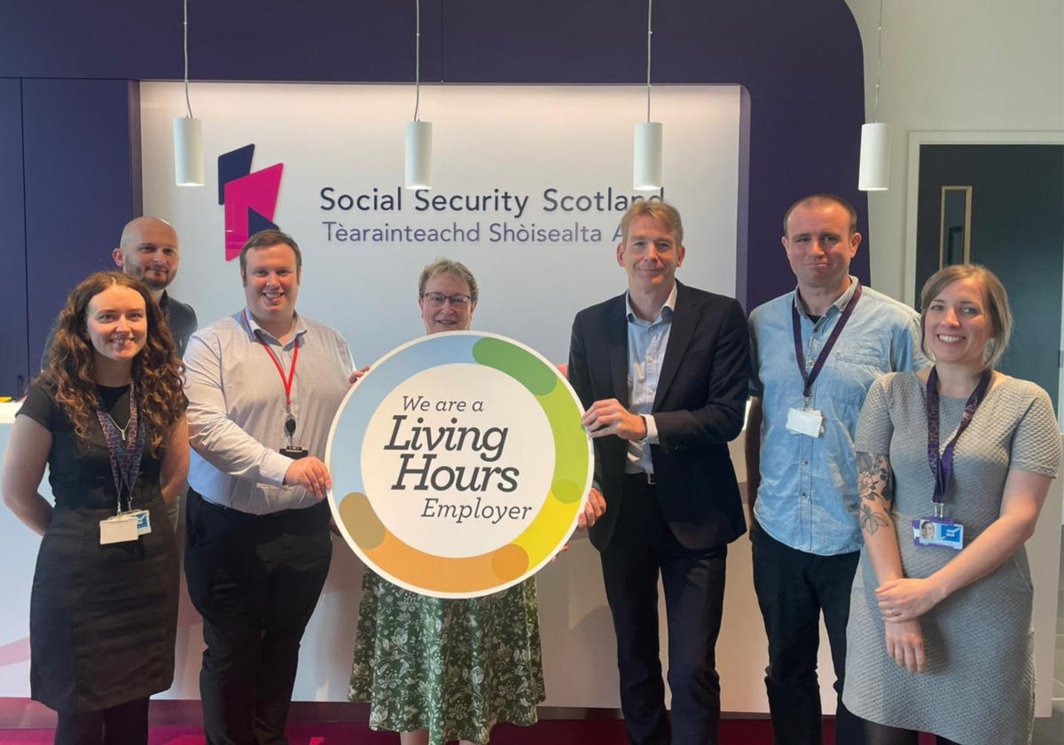 A group of people stand in front of a board which reads 'Social Security Scotland', they are holding a circular placard which states 'we are a Living Hours Employer'